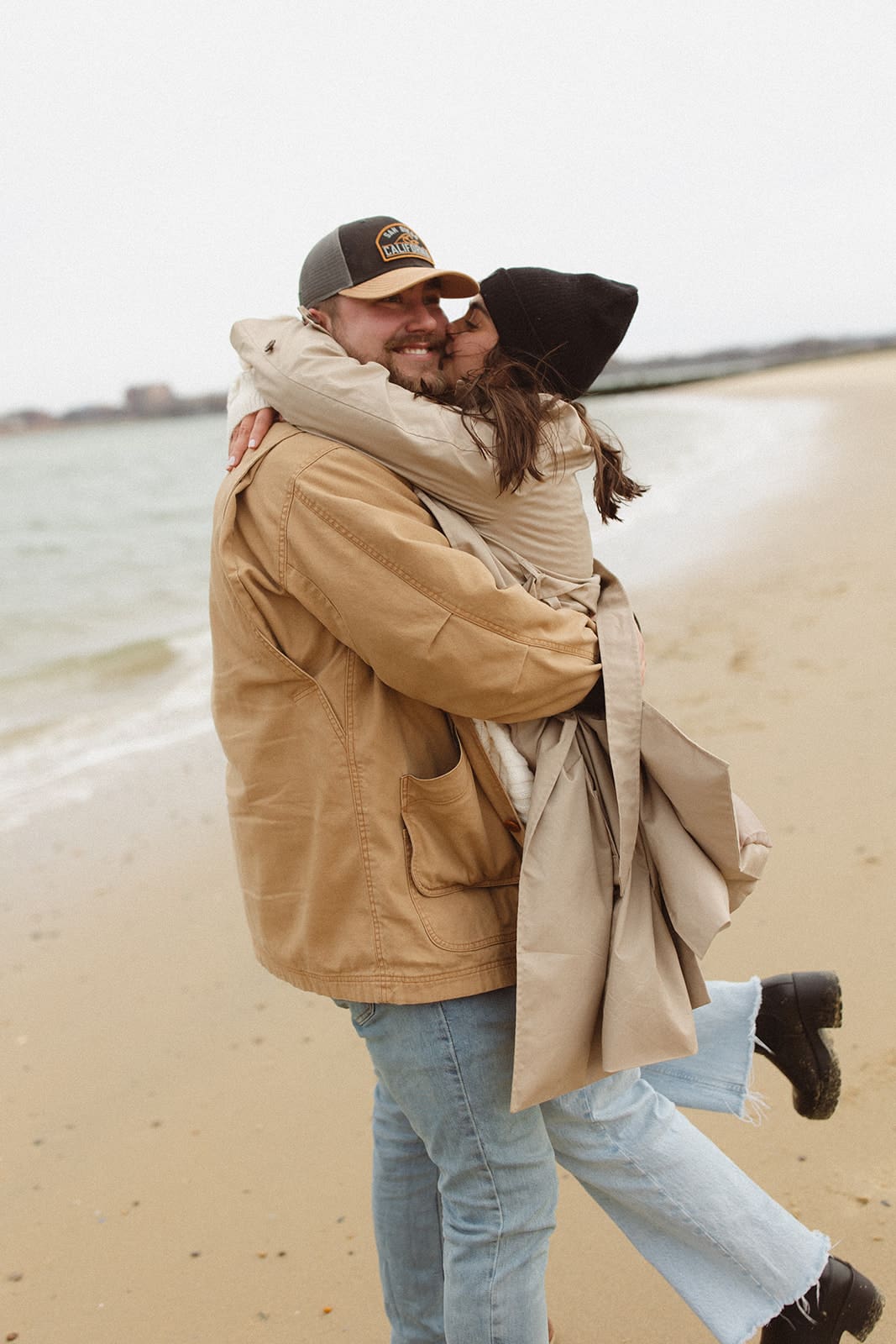 Playful beach moment as a couple hugs and spins near the shoreline, wearing relaxed winter engagement session outfits with neutral coats and denim.
