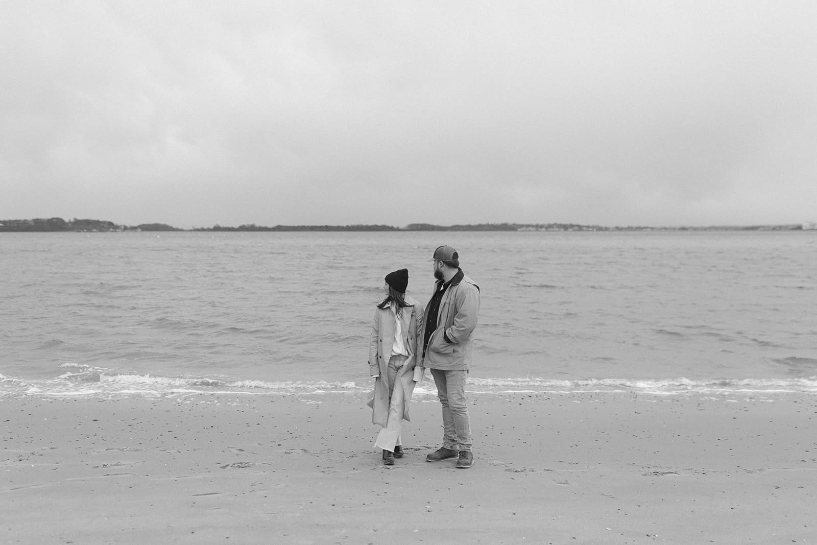 Black and white engagement portrait of a couple standing together by the ocean in relaxed winter engagement session outfits.
