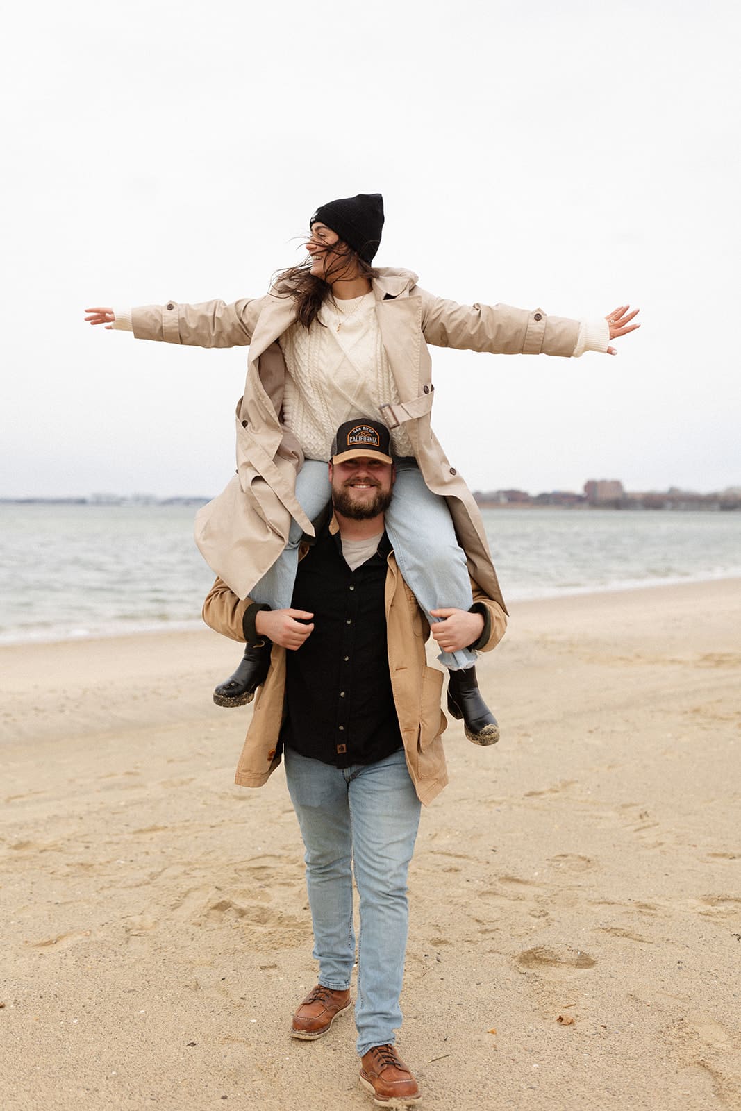 Groom carrying his fiancée on his shoulders along a quiet beach, showing layered winter engagement session outfits with a trench coat and knit beanie.
