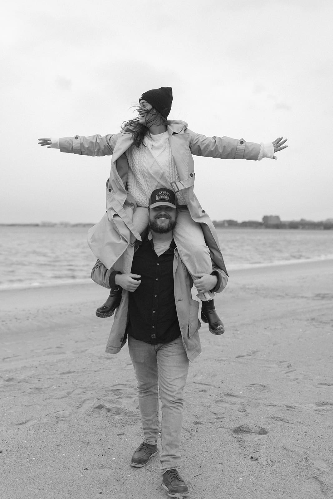 Groom carrying his fiancée on his shoulders along a quiet beach during a playful winter engagement session.
