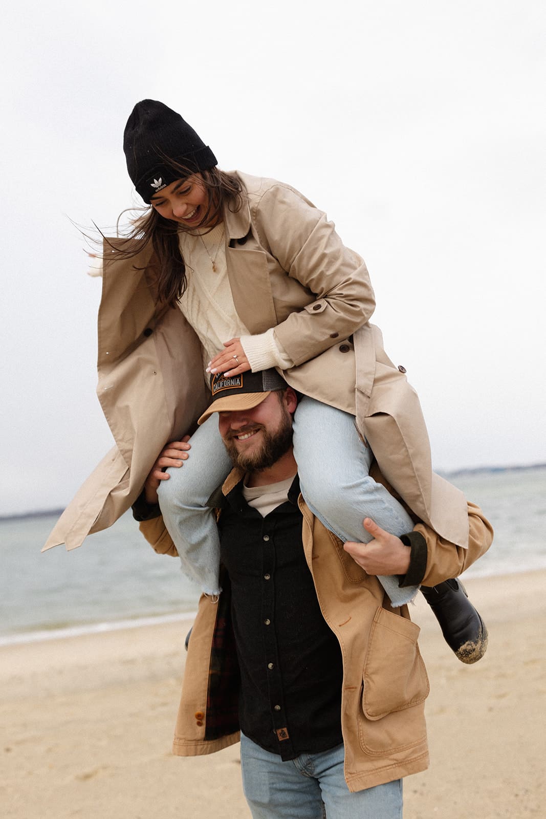 Playful piggyback moment on the beach featuring layered winter engagement session outfits perfect for a cold coastal session.
