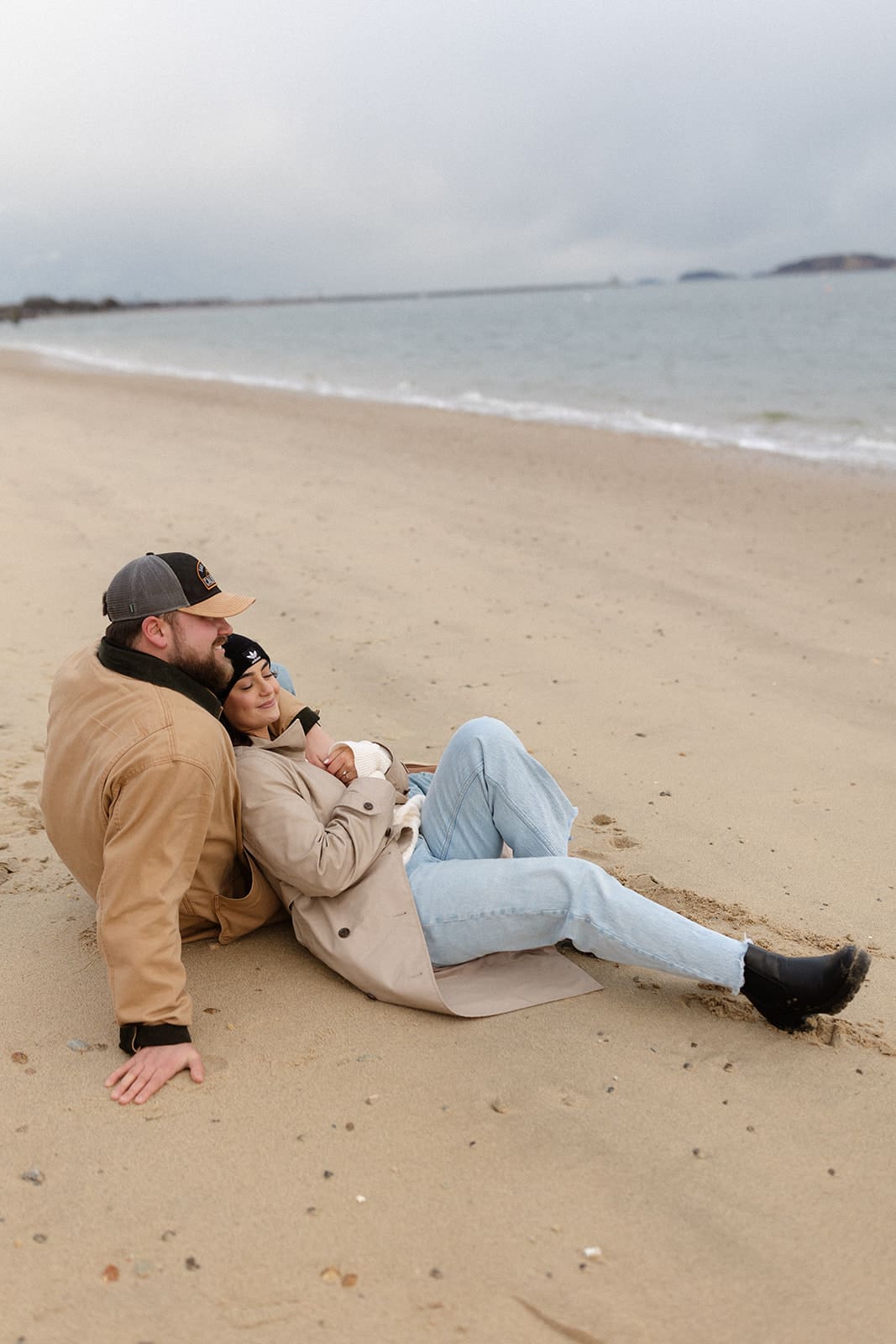 Couple sitting along the shoreline in neutral coats and denim during a peaceful winter engagement session by the water.
