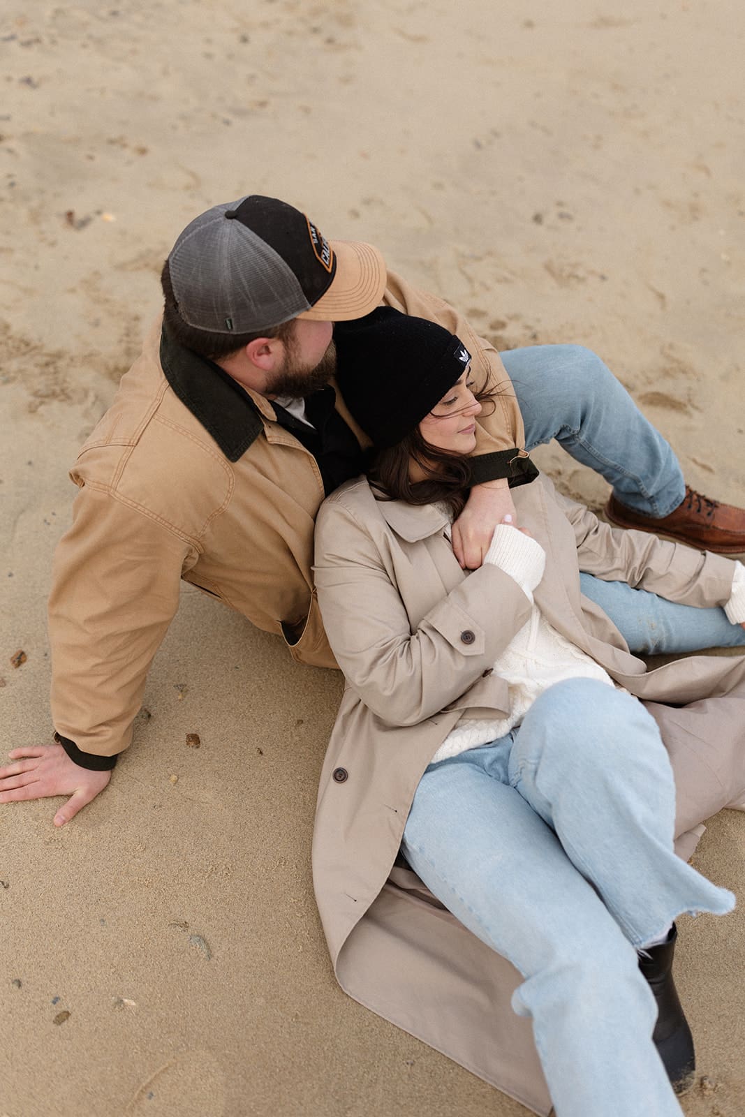 Couple relaxing in the sand wearing neutral winter engagement session outfits including a trench coat, knit beanie, and tan jacket.
