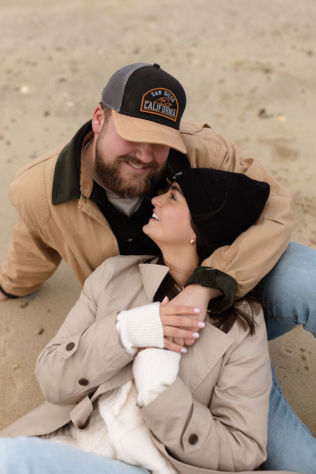 Cozy beach portrait of a couple laughing together while bundled in warm winter engagement session outfits.
