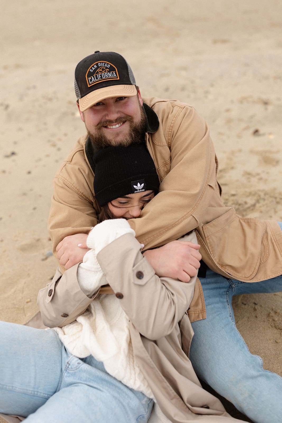 Couple sitting in the sand laughing while bundled in winter engagement session outfits during a relaxed coastal engagement shoot.
