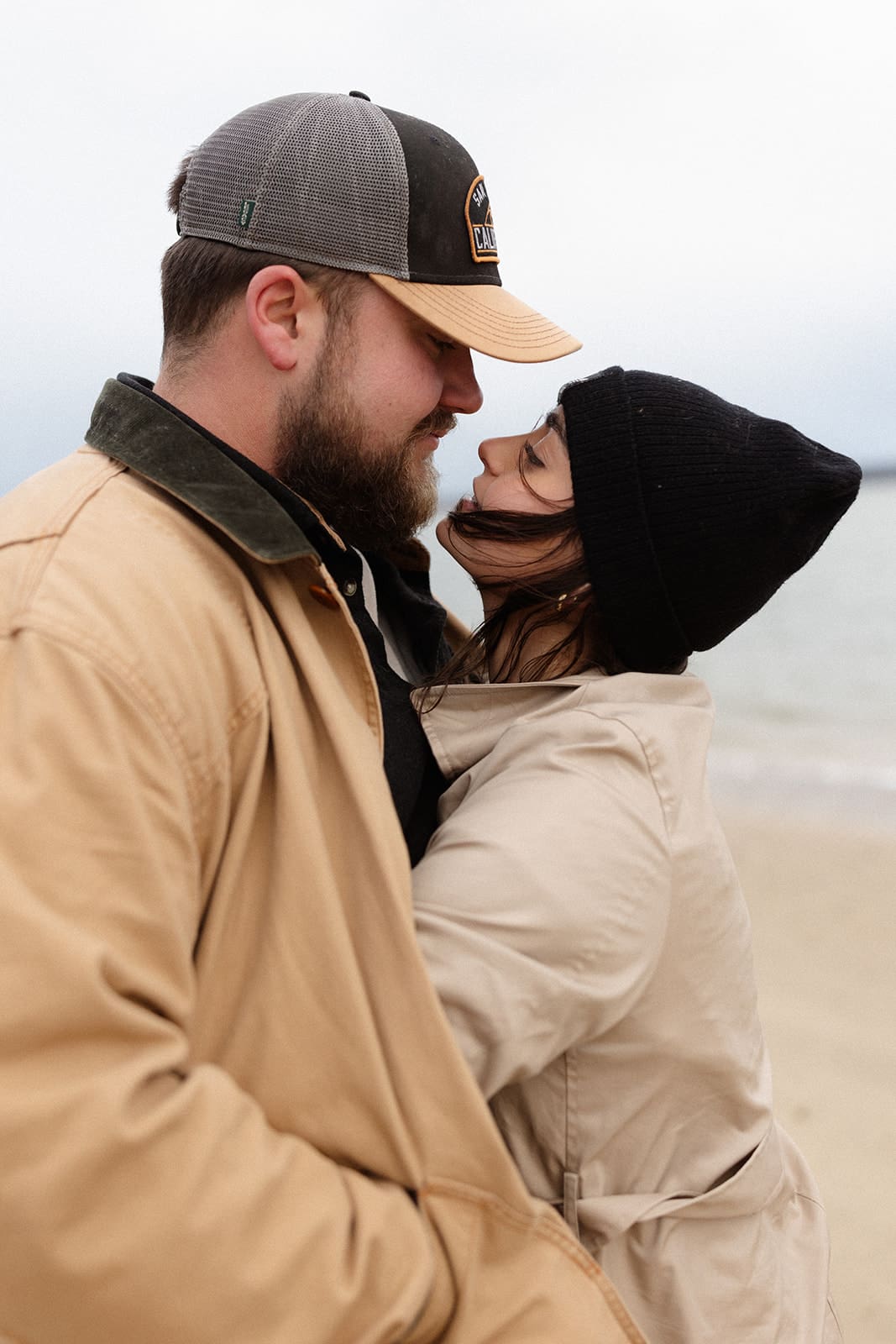 Intimate engagement portrait of a couple leaning together on a breezy shoreline in cozy neutral coats.
