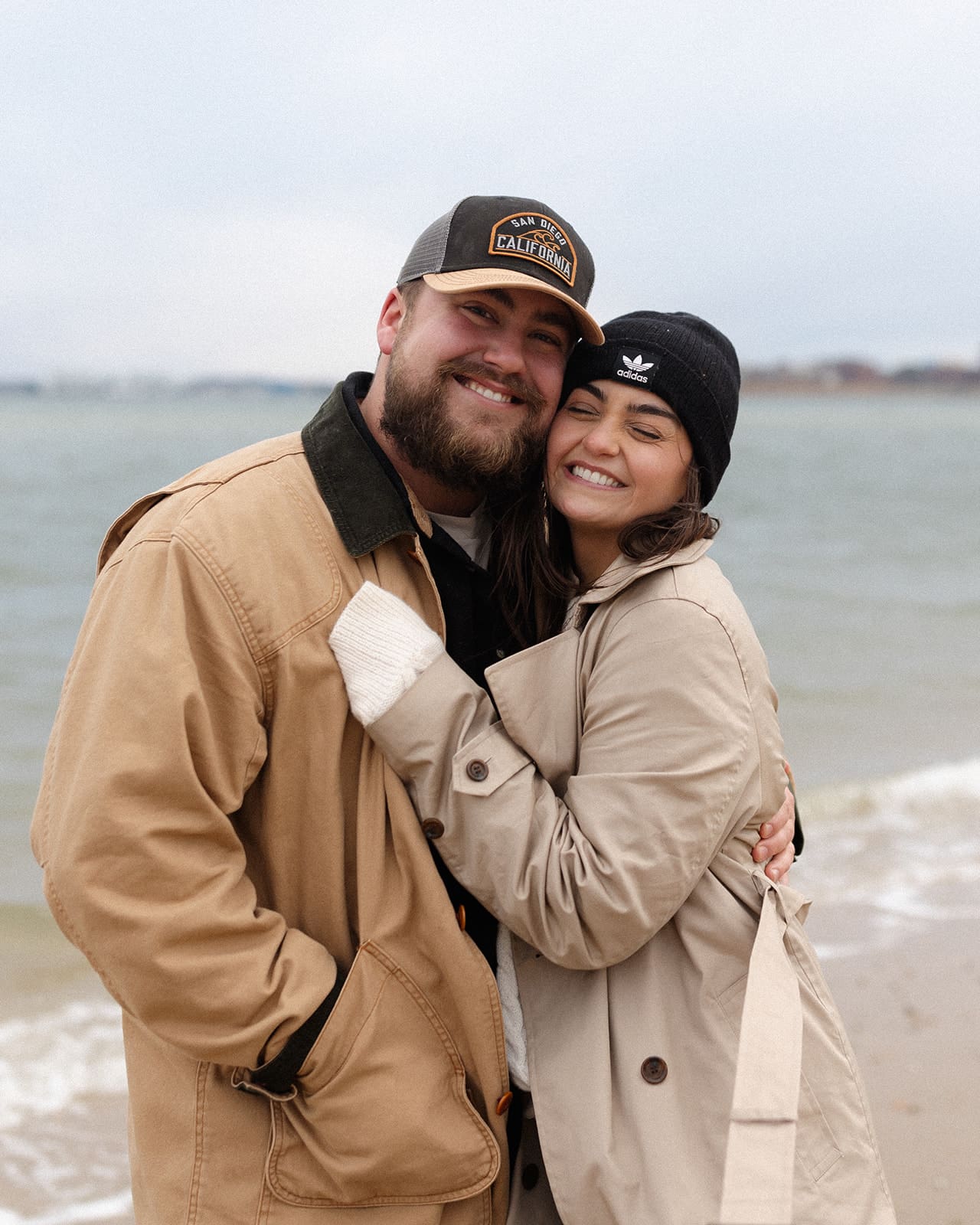 Joyful couple hugging by the water during a winter beach engagement session with soft neutral-toned winter engagement session outfits.

