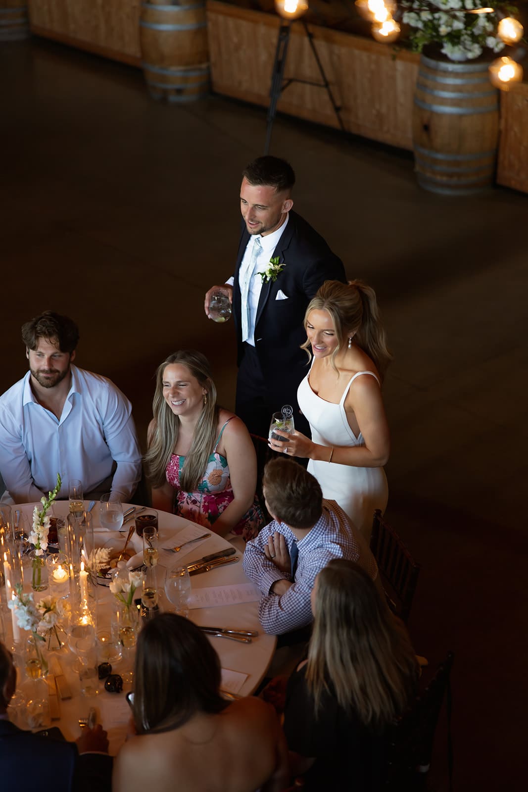Bride and groom standing with friends during reception speeches at a candlelit wedding reception table.