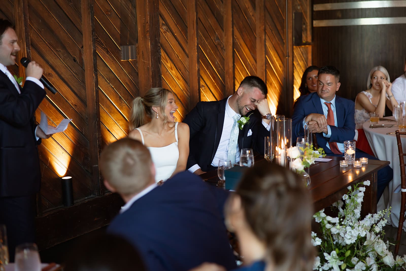 Best man giving a speech during a candlelit barn wedding reception while the bride and groom laugh at the sweetheart table.