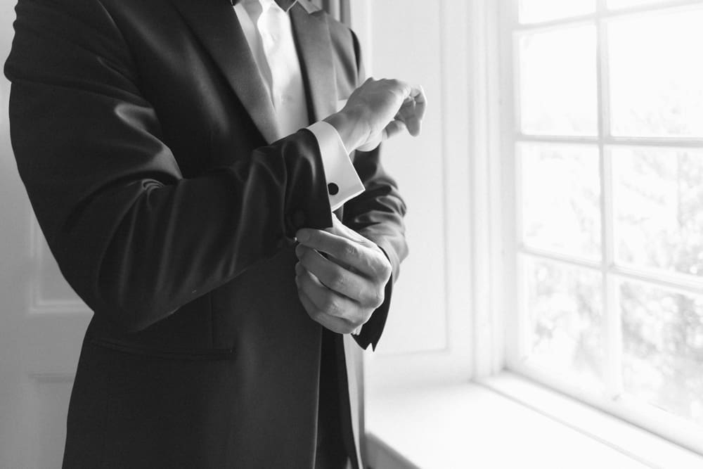 Close-up of groom fastening his cufflink in a black and white portrait by the window

