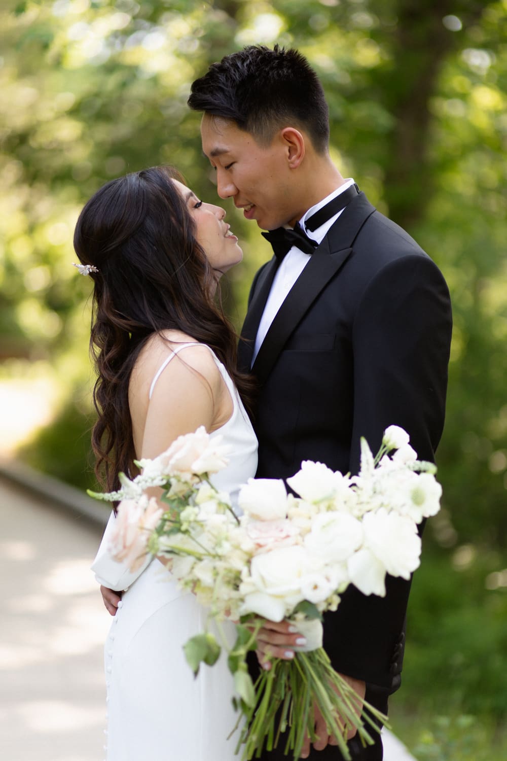 Bride and groom standing close together on a tree-lined path, holding a white and blush bouquet and leaning in softly
