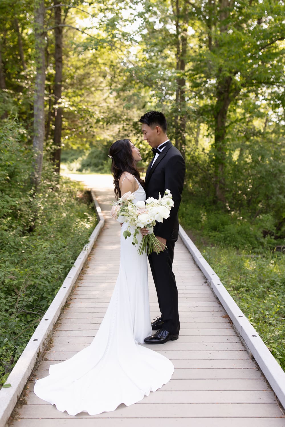 Bride and groom stand together on a wooded path, holding a soft white and blush bouquet in natural light