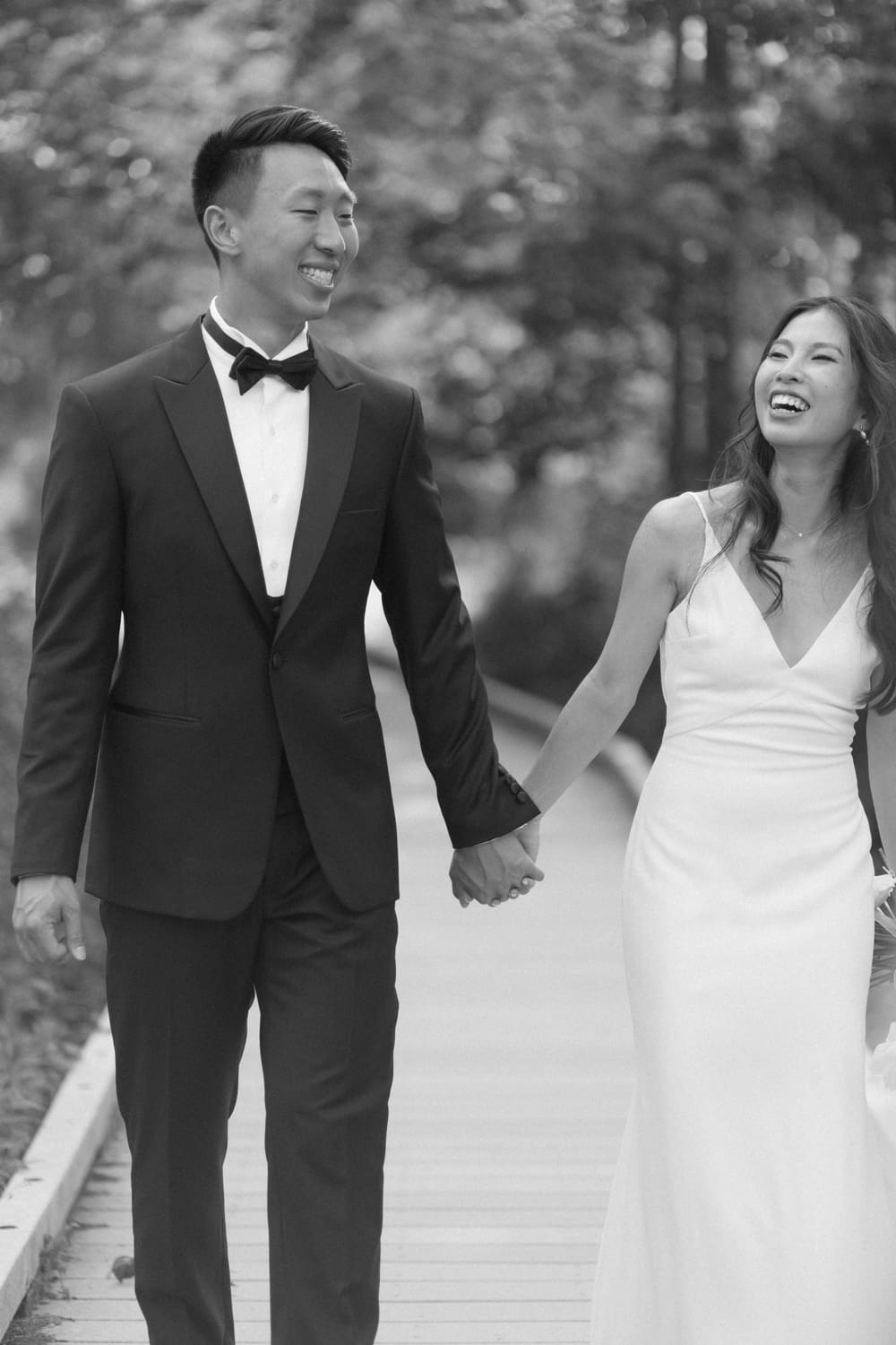 Bride and groom walking hand in hand along a wooded path, smiling at each other in a quiet moment together
