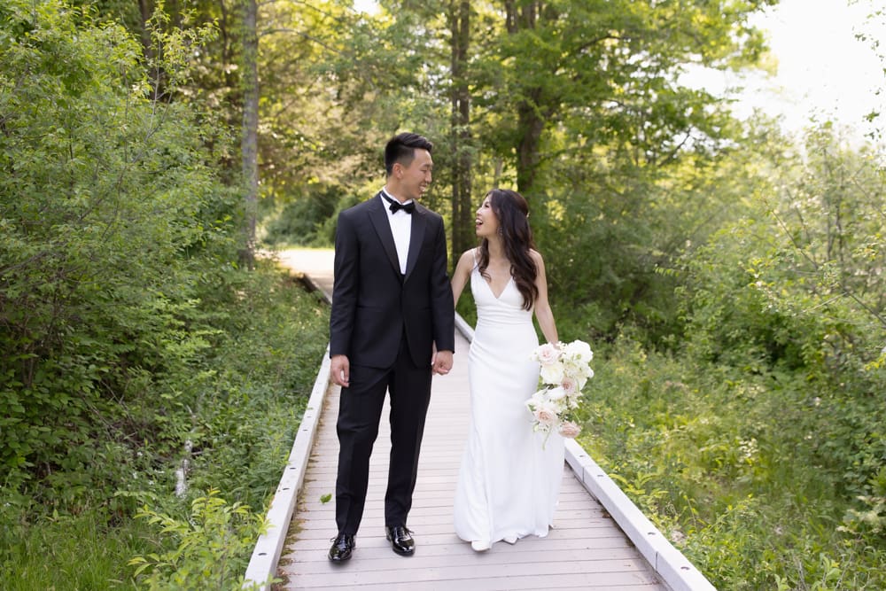 Bride and groom walking along a tree-lined path, holding hands and talking quietly together
