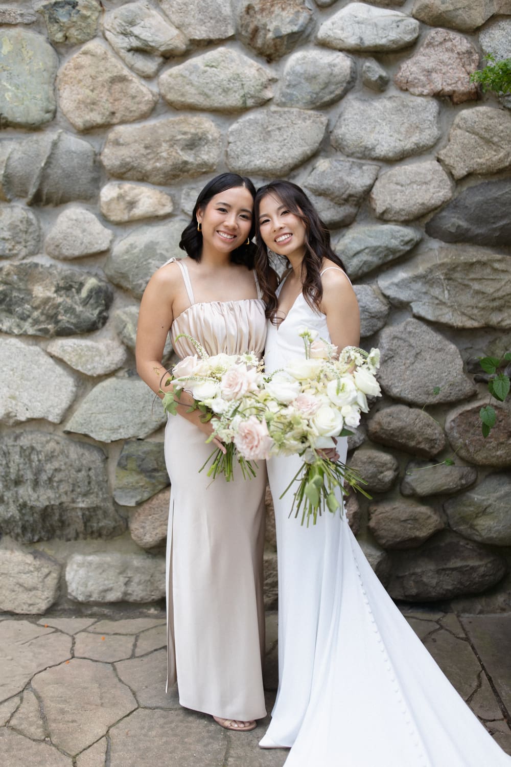 Bride and bridesmaid standing together with bouquets in front of a textured stone wall