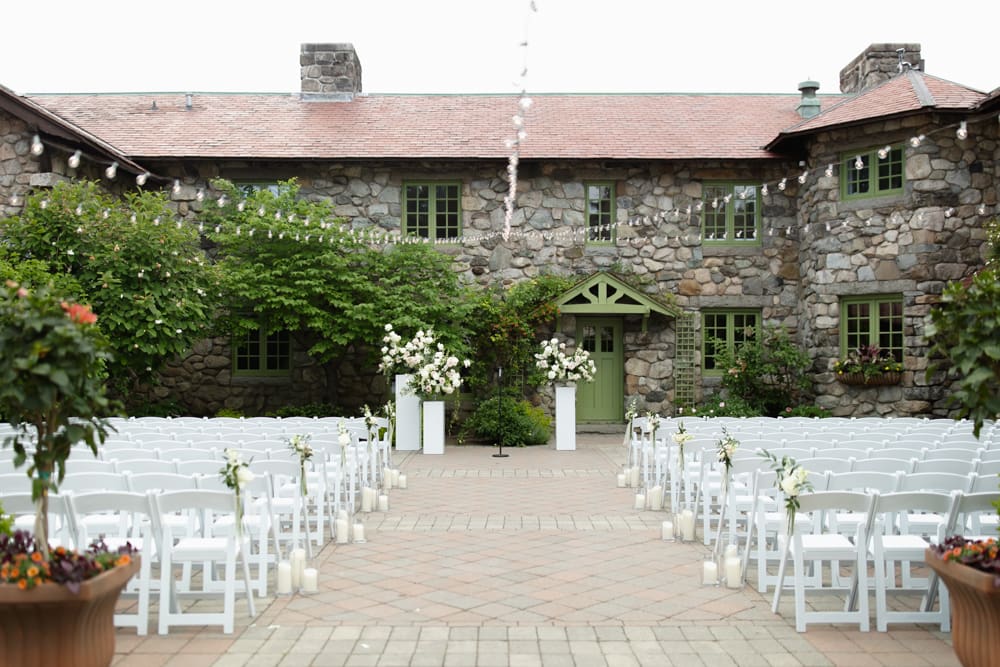 Outdoor ceremony setup with white chairs, candles, and floral arrangements at a Massachusetts wedding venue with a rustic stone facade
