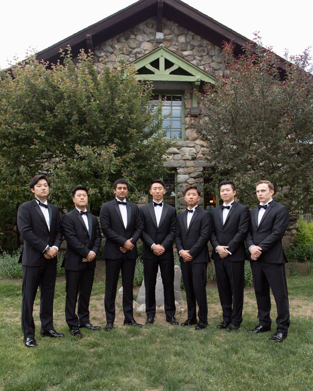 Groom and groomsmen in black tuxedos standing together in front of a stone building at a Massachusetts wedding venue