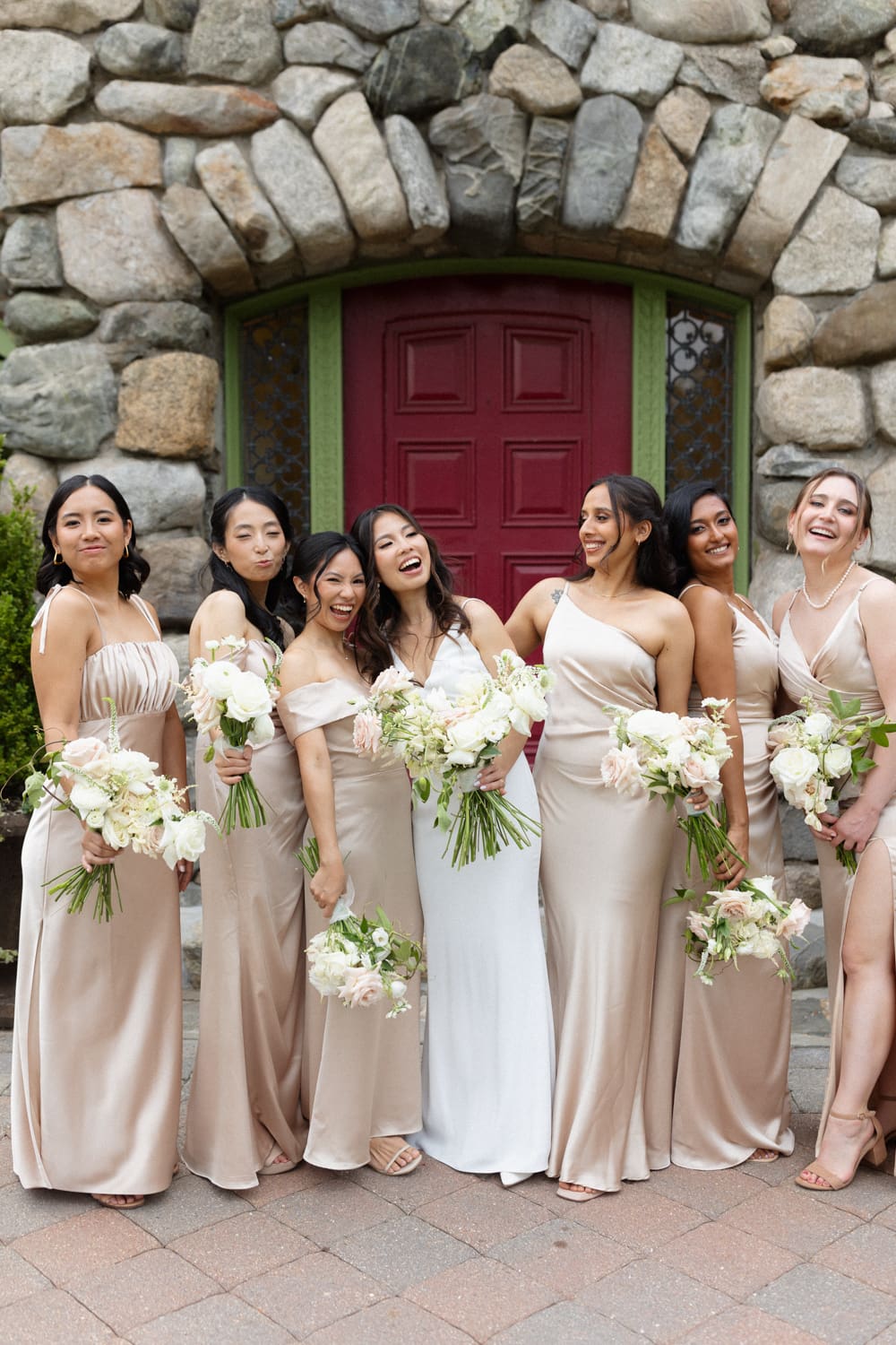 Bride and bridesmaids in neutral satin dresses laughing together in front of a rustic stone doorway