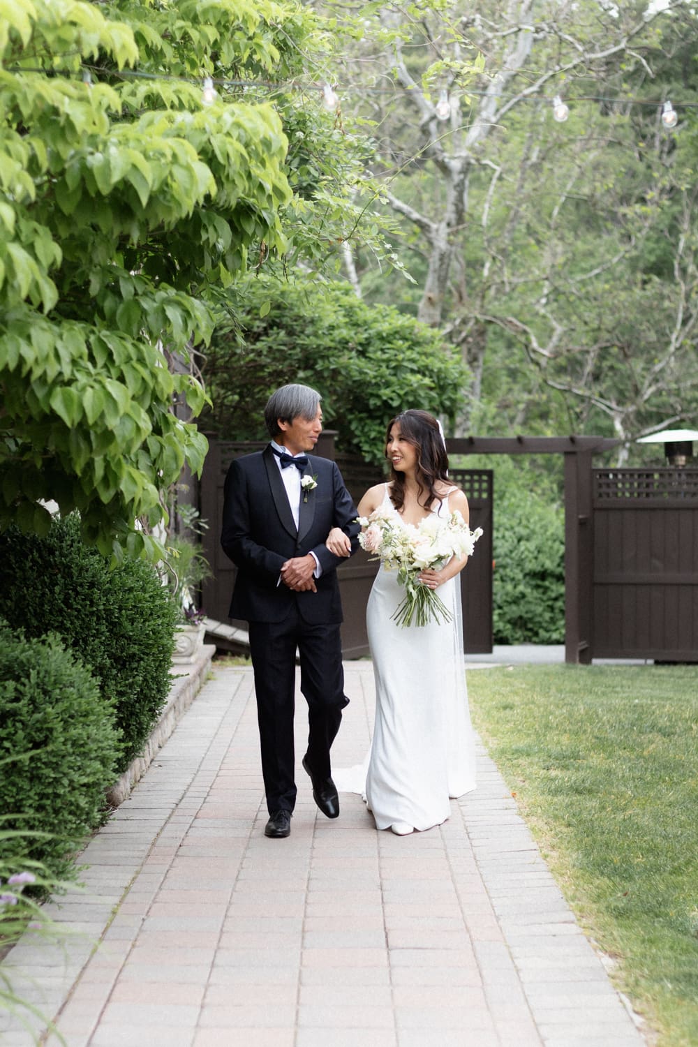 Bride walking arm in arm with her father along a garden path, holding a loose white floral bouquet