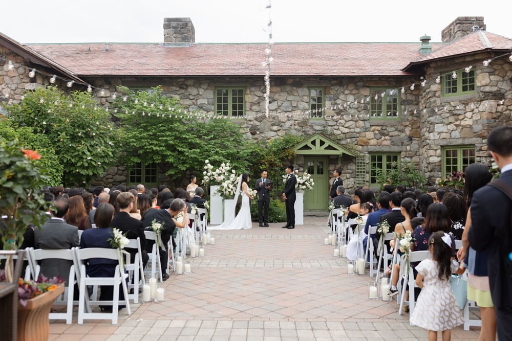 Wide view of an outdoor courtyard ceremony with guests seated and a stone estate backdrop at a Massachusetts wedding venue