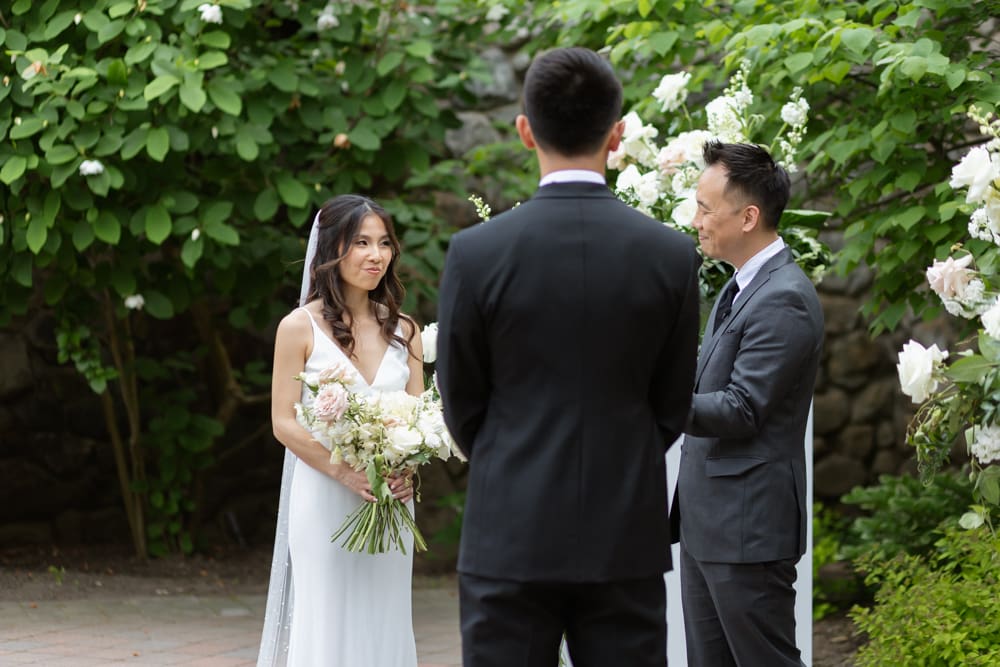 Bride holding her bouquet and listening during the ceremony, facing her partner in a garden setting