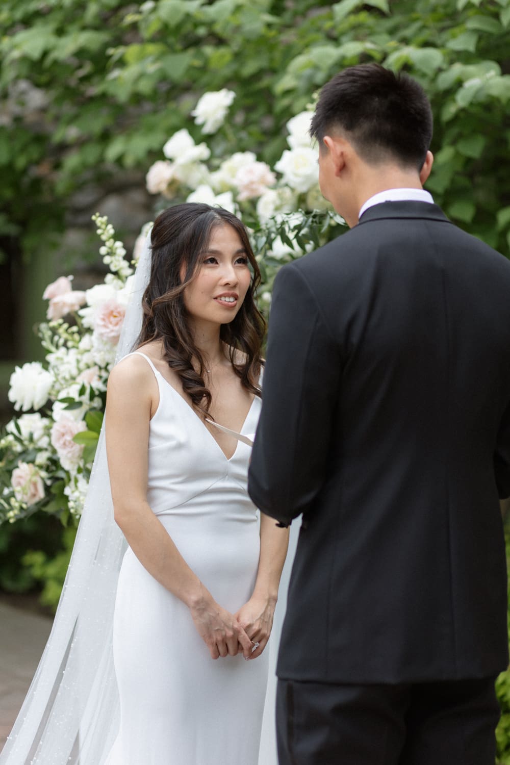 Bride listening closely during vows, holding her bouquet in front of soft white floral arrangements