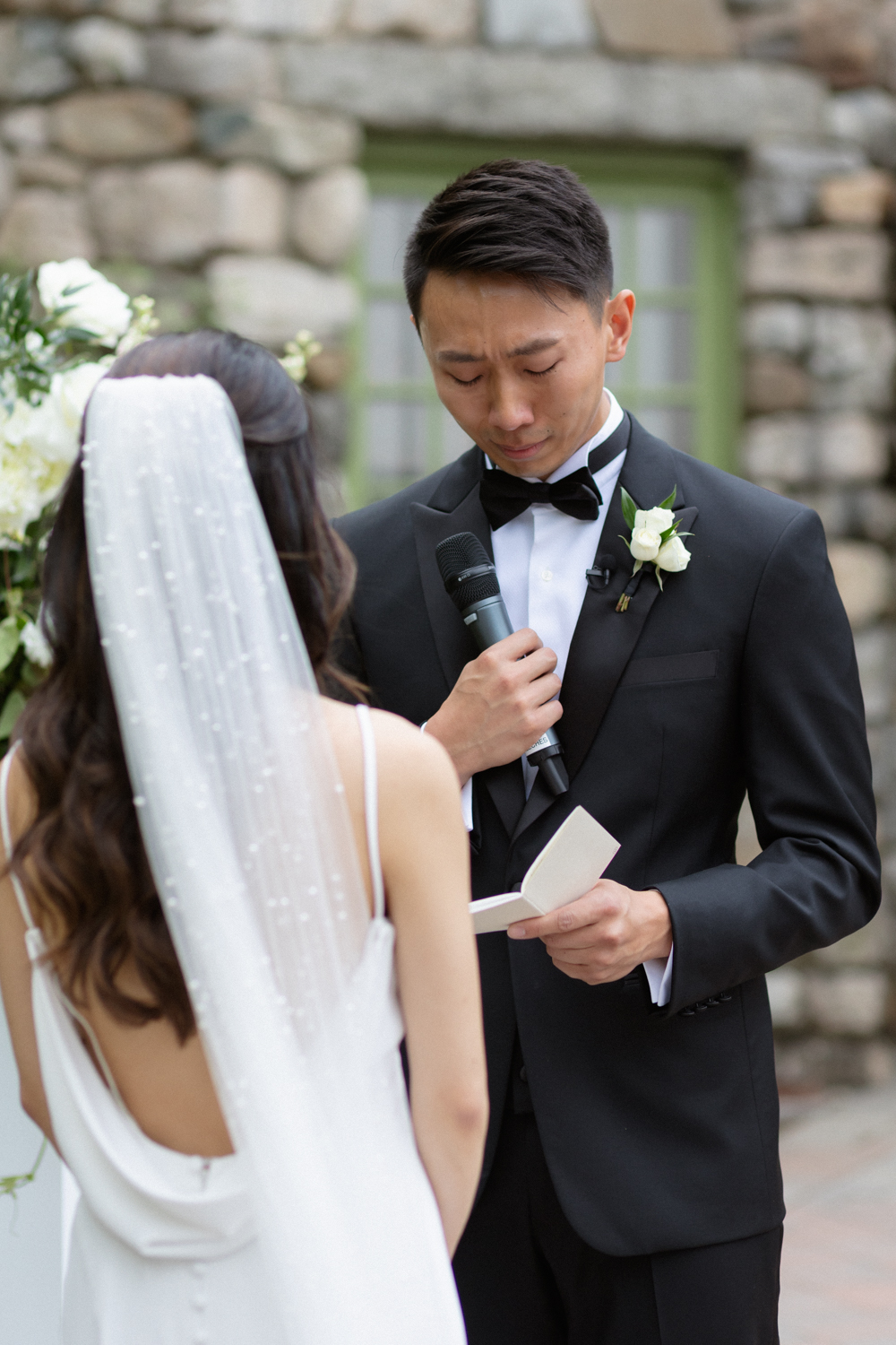 Groom reading his vows while holding a microphone during an outdoor ceremony, with the bride standing close in front of him