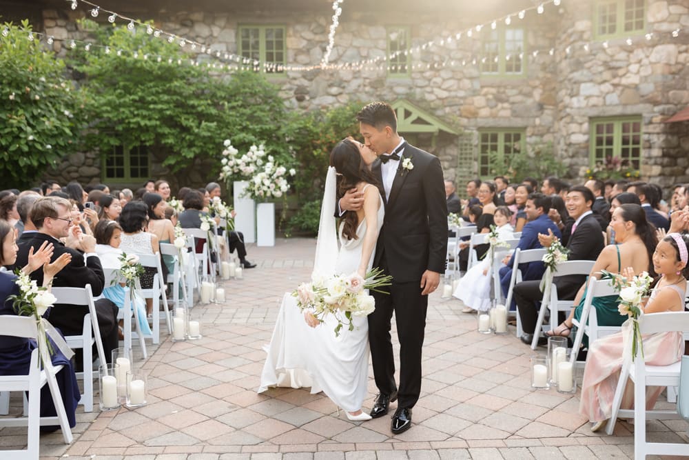Bride and groom share a kiss in a candlelit courtyard ceremony with guests seated under string lights at a Massachusetts wedding venue