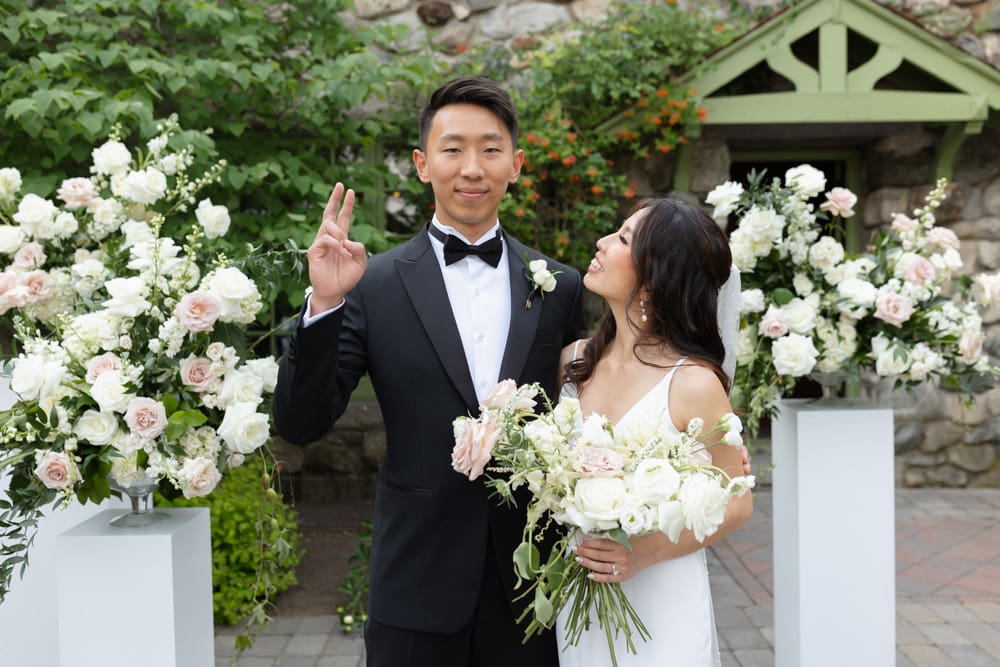 Bride and groom standing together in front of floral arrangements at a Massachusetts wedding venue, smiling and holding a bouquet
