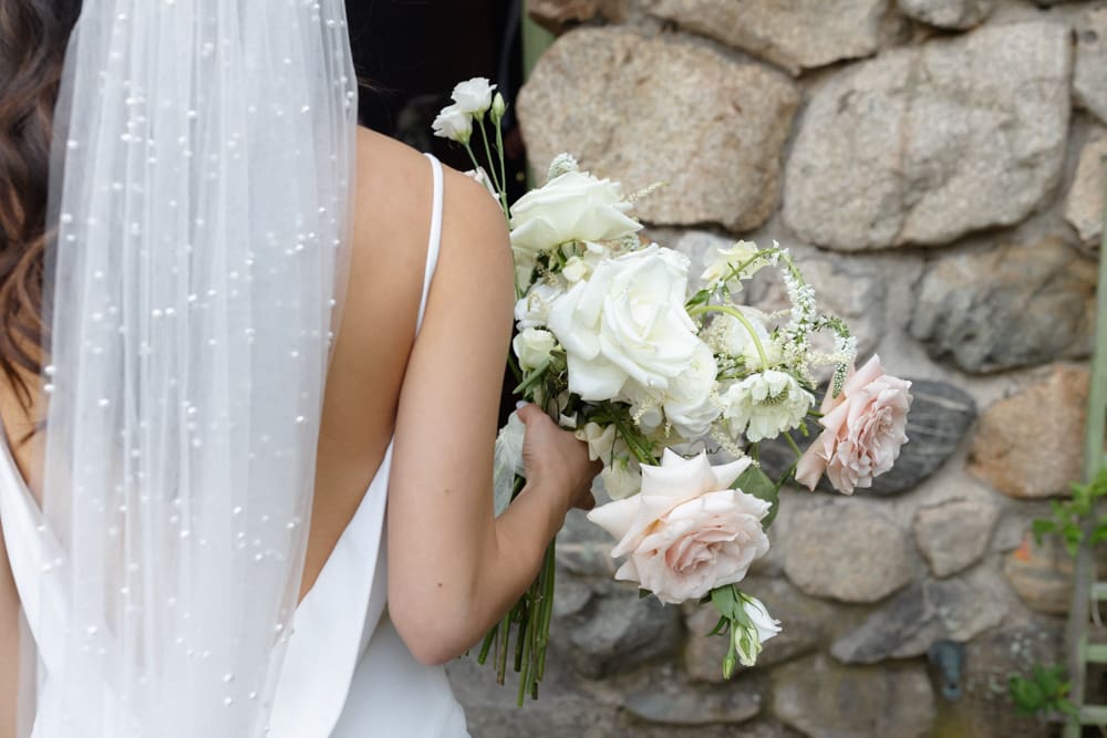 Close-up of bride holding her bouquet with soft blush roses in front of a rustic stone wall

