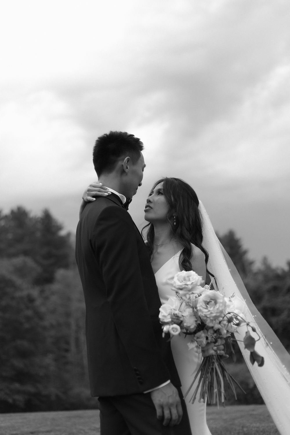 Bride and groom embracing on a grassy field under an overcast sky, holding a soft white and blush bouquet
