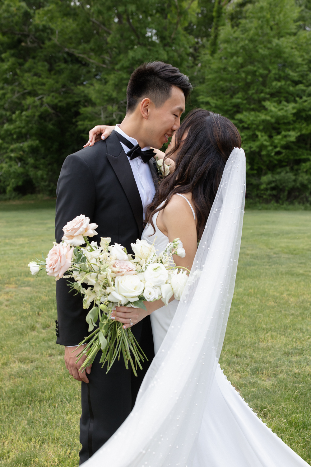 Bride and groom embracing on a grassy field, holding a soft white and blush bouquet and leaning into each other
