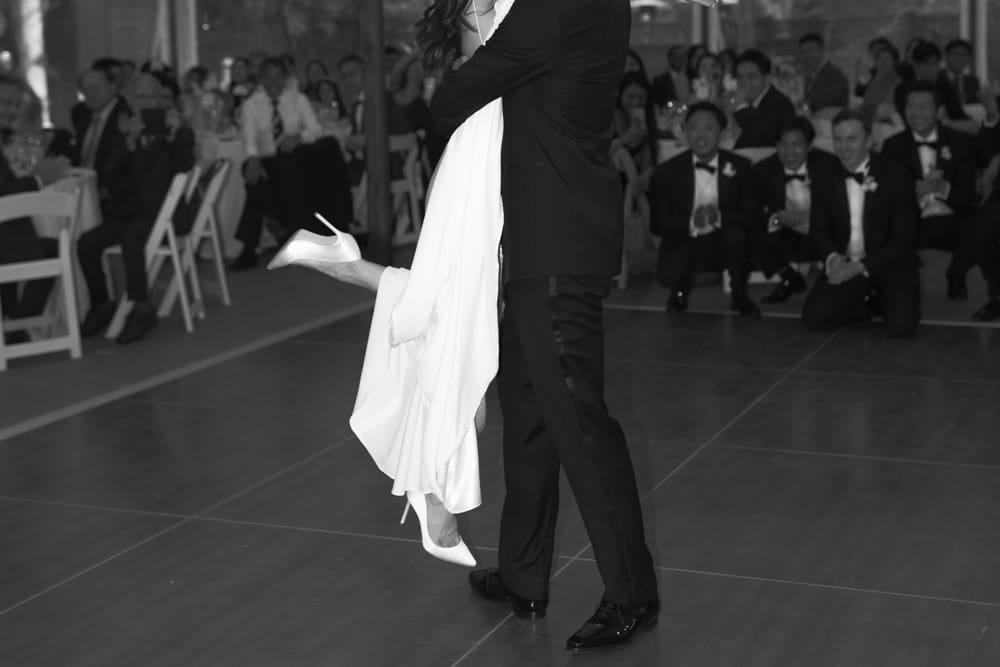 Black and white photo of groom lifting the bride during their first dance while guests watch and smile
