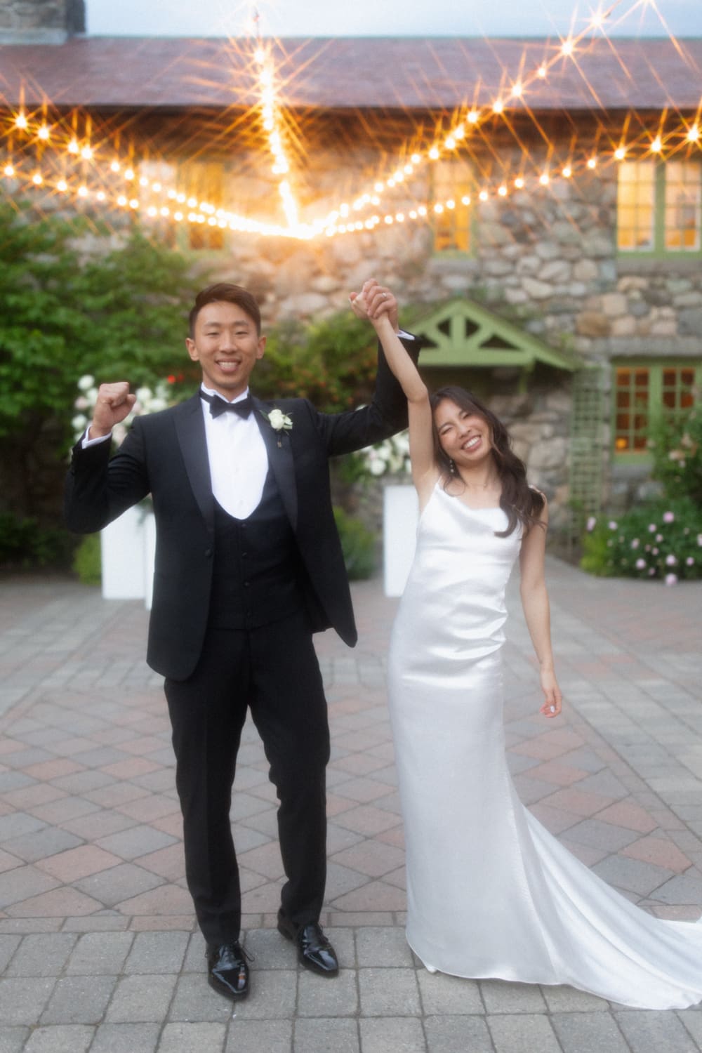 Bride and groom celebrating with hands raised under string lights in a courtyard at a Massachusetts wedding venue