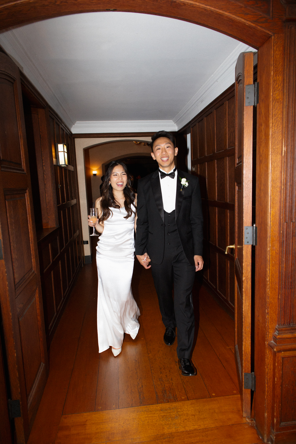 Bride and groom walking through a wood-paneled hallway together, holding hands and smiling after the reception at a Massachusetts wedding venue