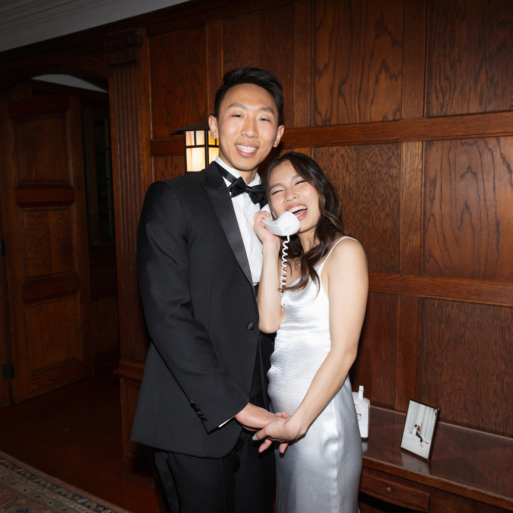 Bride and groom smiling together indoors, holding a vintage phone in a wood-paneled room