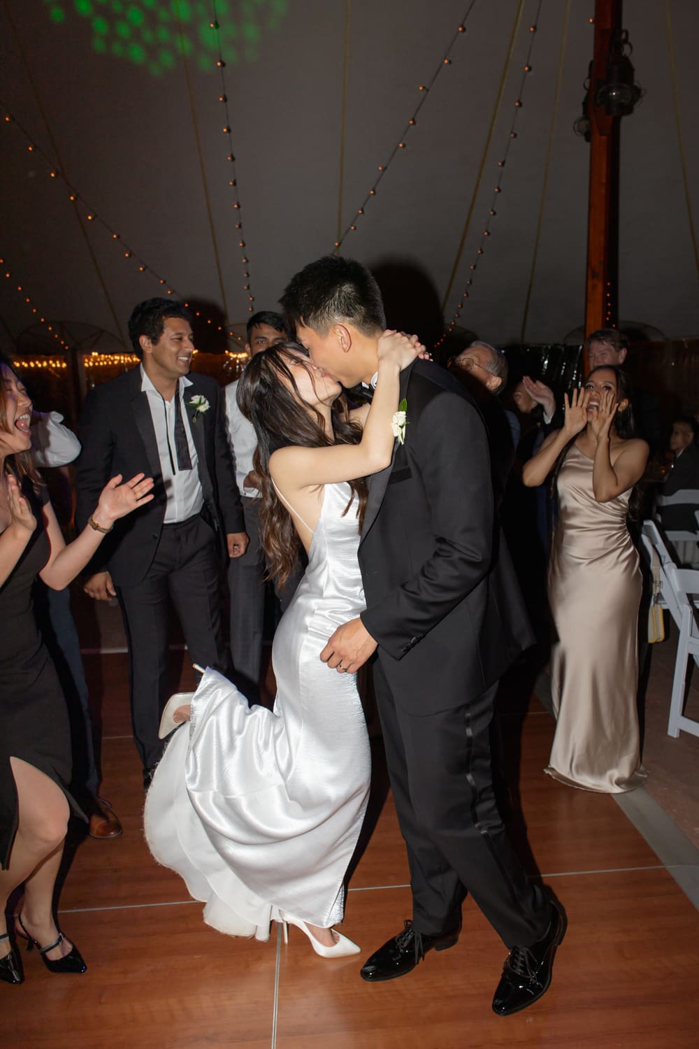 Bride and groom kissing on a packed dance floor surrounded by cheering guests
