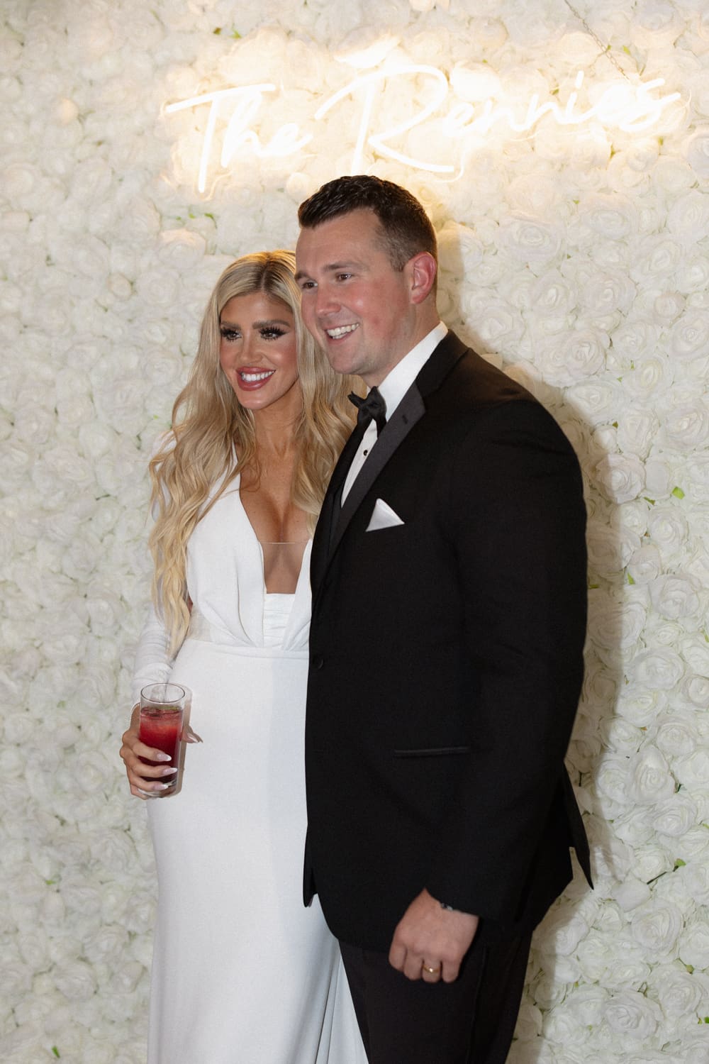Bride and groom smiling in front of a white floral wall with a neon sign during their reception.