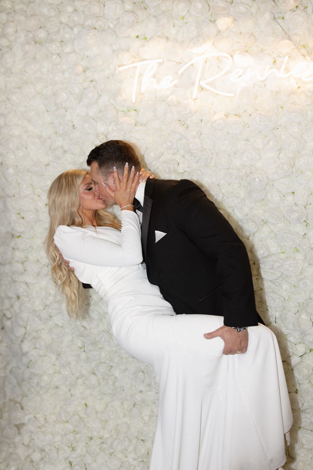 Bride and groom kissing in front of a white rose backdrop with soft lighting at a wedding venue in Rhode Island.