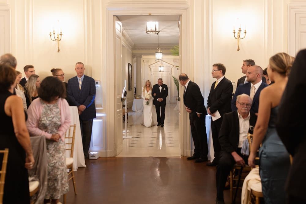 Bride walking through a grand hallway with her father just before the ceremony, guests waiting quietly inside.
