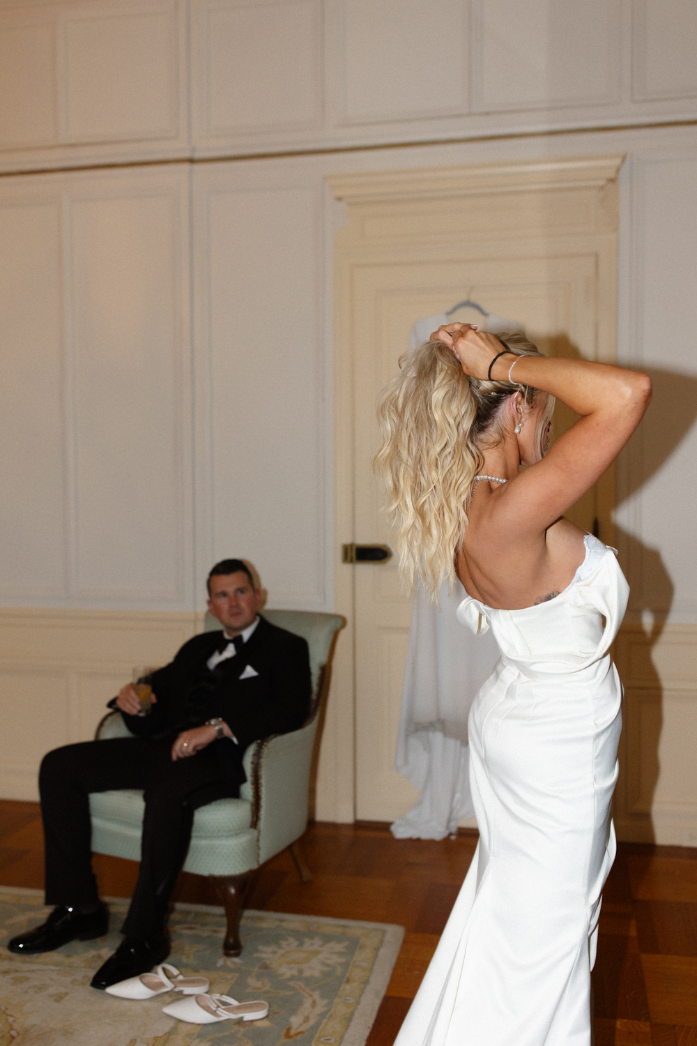 Bride adjusting her hair in a fitted white gown while the groom sits nearby watching in a quiet moment at a wedding venue in Rhode Island.