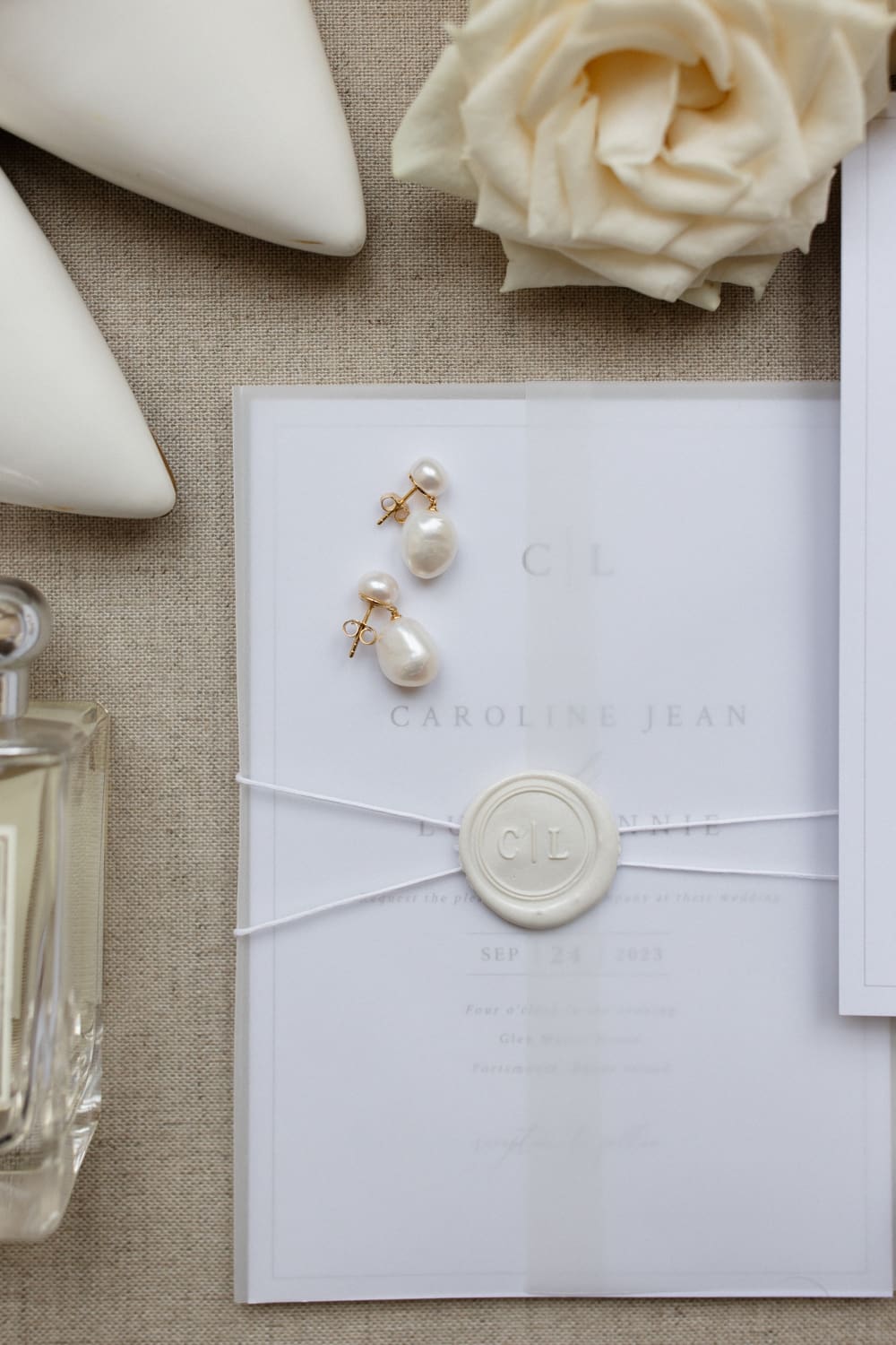 Wedding invitation suite with pearl earrings, perfume, and neutral details styled on soft linen.
