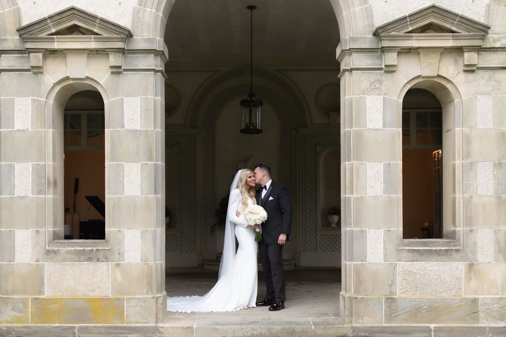 Bride and groom standing together beneath a grand stone archway outside a wedding venue in Rhode Island.