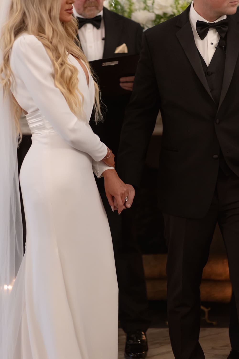Bride and groom holding hands during an intimate indoor ceremony, framed tightly on their hands and classic attire.
