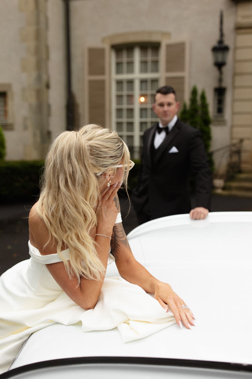 Bride sitting on the hood of a vintage car with the groom just behind her, sharing a relaxed moment outside a wedding venue in Rhode Island.

