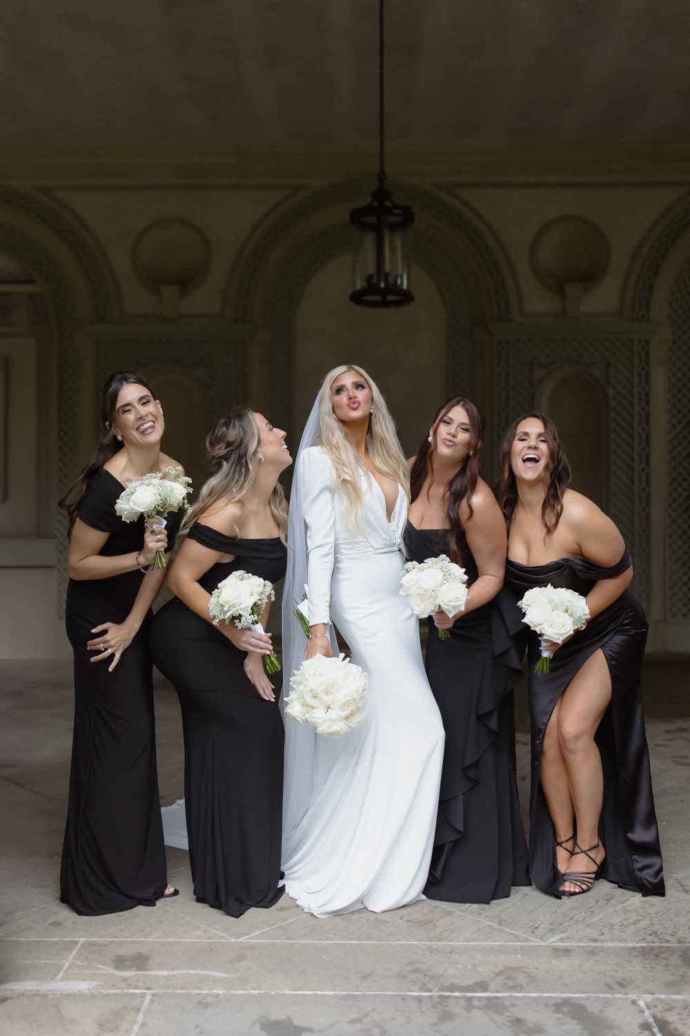 Bride with her bridesmaids in black dresses, holding white bouquets and laughing together beneath a covered courtyard.
