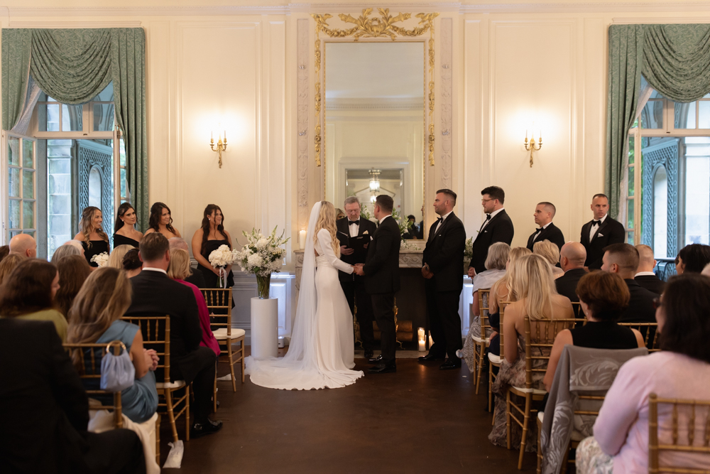 Wide view of an indoor ceremony with bride and groom at the altar surrounded by guests at a wedding venue in Rhode Island.