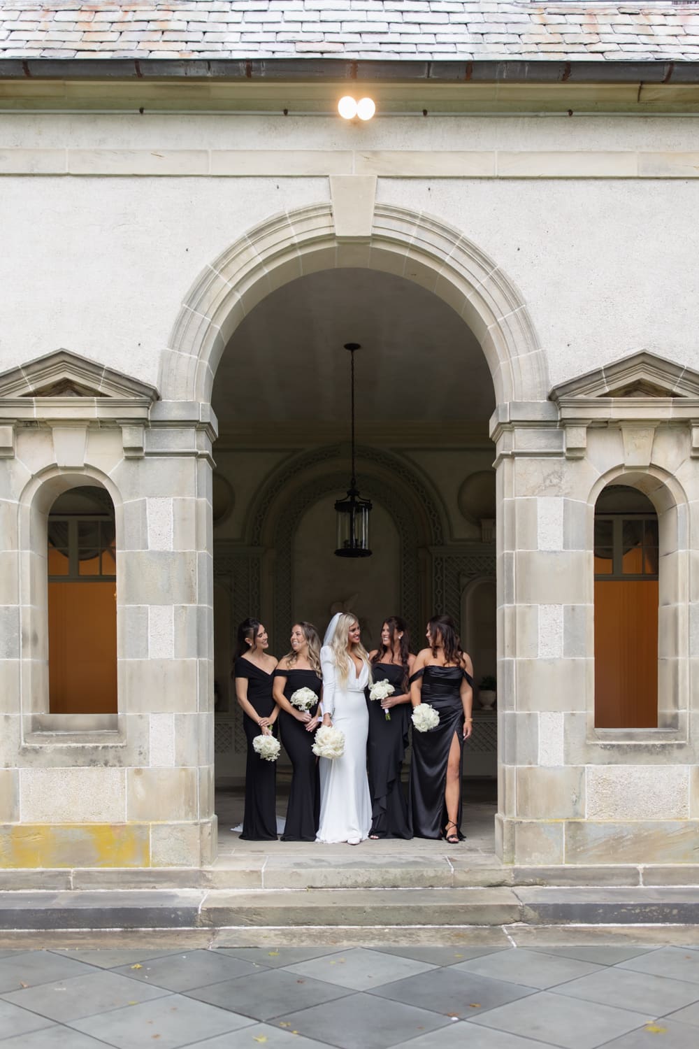 Bride and bridesmaids in black dresses standing together beneath a stone archway outside a wedding venue in Rhode Island.