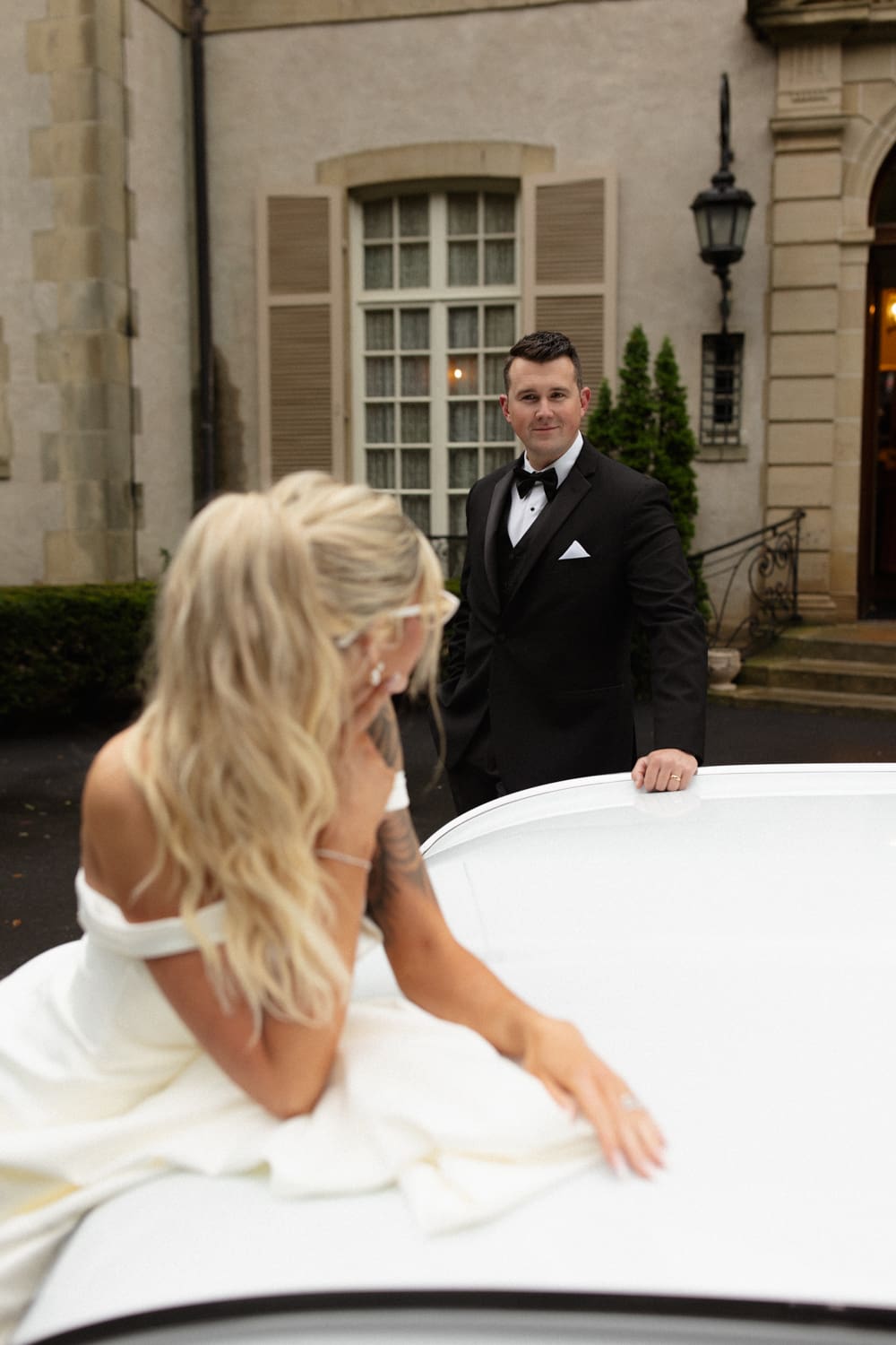Bride sitting on the hood of a classic car while the groom stands nearby outside a wedding venue in Rhode Island.