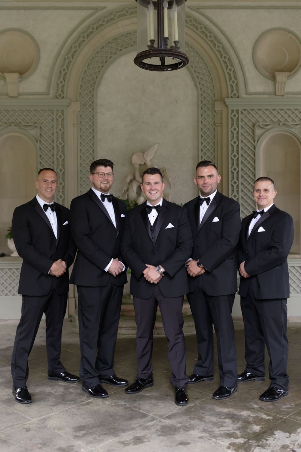 Groomsmen in black tuxedos standing together beneath an arched stone alcove at a wedding venue in Rhode Island.